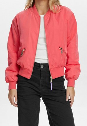 Woman wearing a coral pink bomber jacket over a white shirt and black pants, standing with hands relaxed at sides.