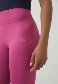 Leggings taille haute fuchsia en tissu texturé, avec une coupe ajustée et un logo argenté sur la cuisse. Une main avec du vernis à ongles naturel est visible.