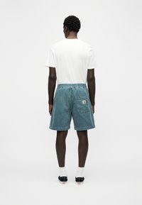 Άνδρας που στέκεται πλάτη, φοράει λευκό t-shirt, μπλε βερμούδα από κορντόρι με πίσω τσέπη, λευκές κάλτσες και μαύρα παπούτσια χωρίς κορδόνια.