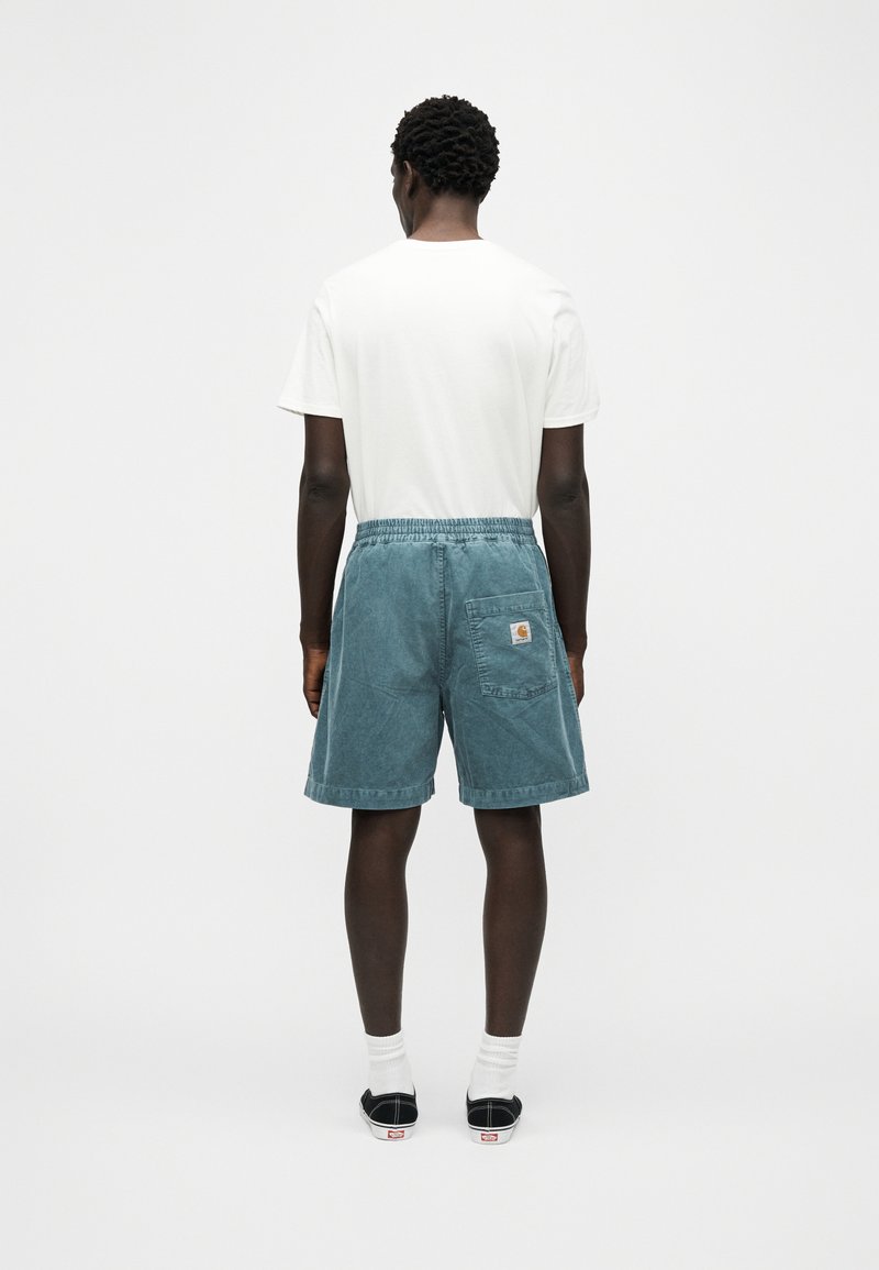 Homme debout de dos, portant un t-shirt blanc, un short en velours côtelé bleu avec poche arrière, des chaussettes blanches et des chaussures noires sans lacets.