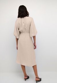 Beige midi mekko, jossa on teksturoitu kangas, pallohihat ja vyötäröltä muotoiltu leikkaus. Takana pieni avainaukko. Mustat sandaalit.