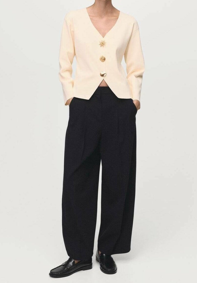 Personne portant un haut crème à manches longues avec un col en V et trois boutons ornés, un pantalon noir taille haute à jambes larges, et des mocassins noirs. Mains dans les poches.
