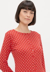 Czerwona bluzka z długim rękawem w białe kropki, z okrągłym dekoltem i zapinana na guziki z przodu. Wykonana z prążkowanej tkaniny o teksturowanym wykończeniu.