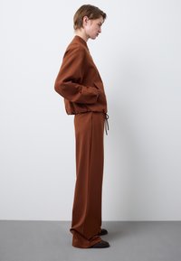 Giacca marrone di taglio ampio con polsini elasticizzati, abbinata a pantaloni a gamba larga coordinati e calzature scure con texture. Materiale liscio e design minimale.