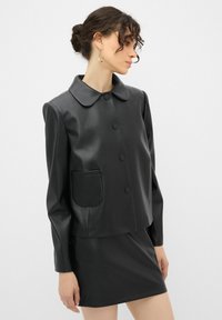 Schwarze Kunstlederjacke mit cropped Design, großem Kragen, Vorderknöpfen und einer aufgesetzten Tasche auf der linken Seite. Glatte Textur.