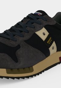 Gros plan d'une basket noire et grise avec des lacets bleu marine, une semelle beige et une bande blanche avec le logo Blauer U.S.A. sur le côté.