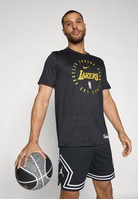 Un t-shirt de sport noir avec un logo "Lakers" jaune, associé à un short noir avec des accents blancs et un ballon de basket texturé à la main.