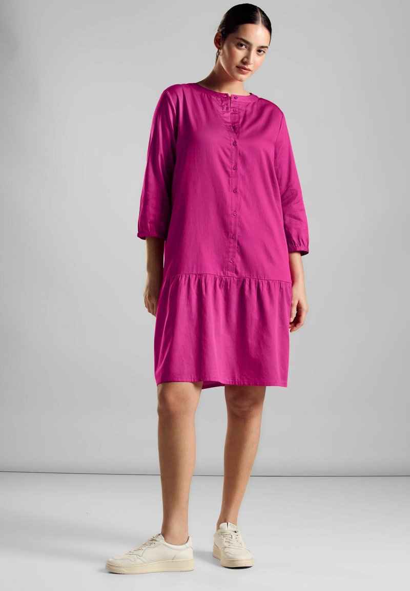 Fuchsia Knopf-up Kleid mit Dreiviertelärmel, einem gestuften Saum und einer lockeren Passform, aus leichtem Stoff gefertigt. Kombiniert mit cremefarbenen Sneakers.