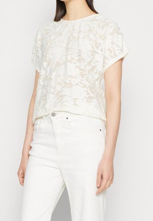 Femme portant un jean blanc taille haute et un chemisier blanc transparent à manches courtes avec un motif floral sur un fond uni.