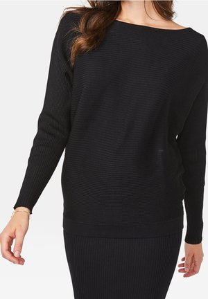 Femme portant un pull côtelé noir à manches longues et une jupe côtelée assortie, debout, les bras détendus le long du corps.