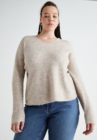 Beige ribbed sweater with a V-neck, long sleeves, and a relaxed fit, paired with blue jeans, against a plain background.