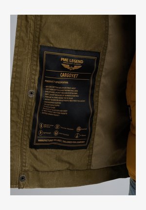 La giacca in misto cotone e nylon verde presenta un'etichetta nera con testo dorato, che riporta specifiche e istruzioni per la cura all'interno.