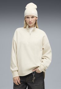Cremfarbener Pullover mit Viertelreißverschluss, Waffelstruktur, mit dezentem Logo und weiten Ärmeln, kombiniert mit einer passenden Mütze. Schlüssel an der Hose befestigt.