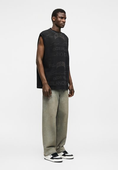 Νεαρός άνδρας που στέκεται, φοράει αμάνικο μαύρο γραφικό πουκάμισο, άνετο ξεθωριασμένο γκρι τζιν και ασπρόμαυρα αθλητικά παπούτσια, με φόντο απλό λευκό.