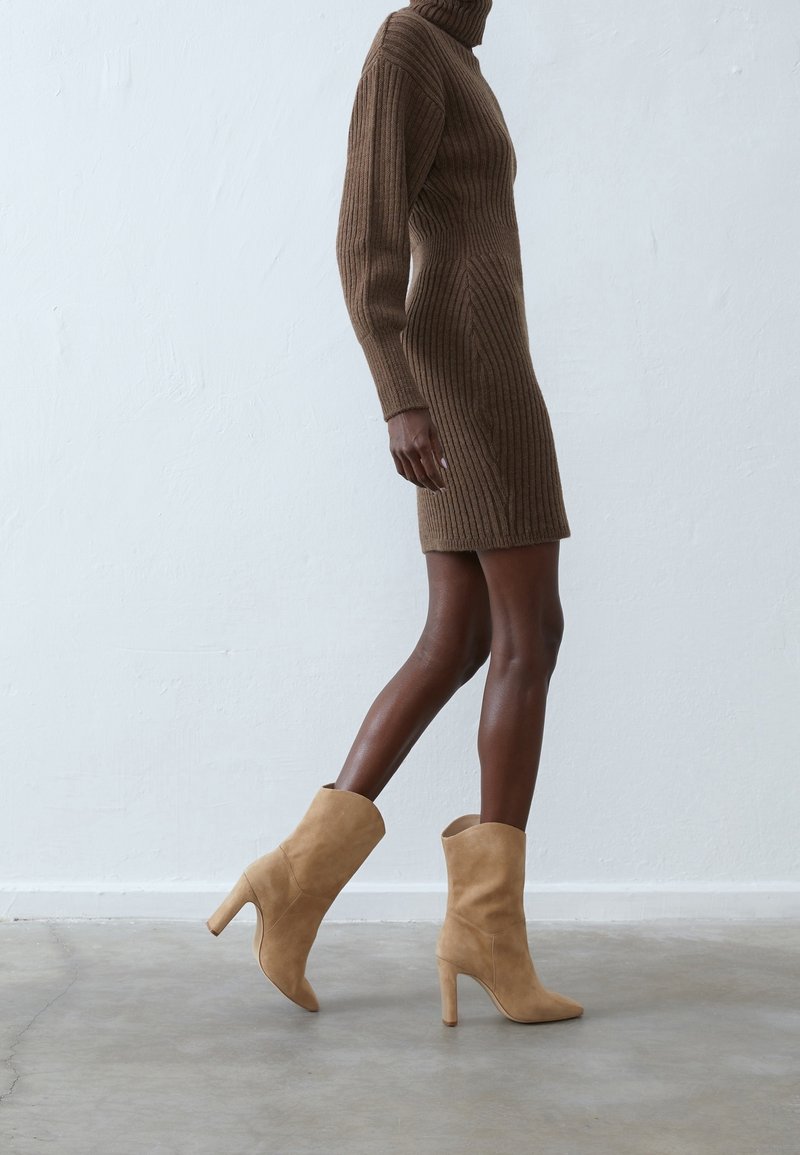 Femme marchant avec une robe en maille côtelée marron et des bottines beiges en daim à talons hauts sur un sol en béton gris.