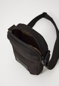 Borsa a tracolla nera in tessuto con chiusura a zip, esterno testurizzato, tracolla regolabile e foderatura interna marrone. Patch del logo visibile sul davanti.