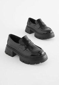 Schwarze Lacklederloafer mit einer klobigen Gummisohle, die einen dekorativen Riemen auf der Vorderseite und eine glatte, glänzende Oberfläche aufweisen.