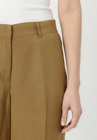 Pantalones de lino marrón de pierna ancha con una textura suave; cuentan con un cierre de botón y gancho, trabillas para cinturón y bolsillos laterales.