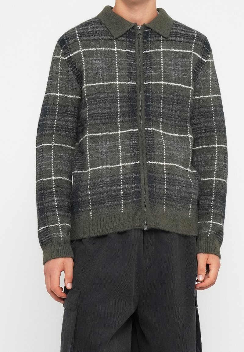Personne portant un pull zippé vert foncé et gris avec un motif à carreaux blancs et un pantalon foncé avec des poches latérales.
