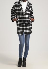 Blazer oversize a quadri in bianco e nero con tessuto spesso, rever a lancia e due tasche frontali, abbinato a jeans skinny blu e stivaletti alla caviglia.