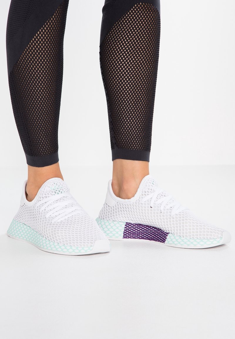 White mesh athletic shoes feature a light turquoise sole with a purple accent on the side, showcasing a breathable design and textured surface.
