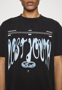 Camiseta de algodón negra con un diseño abstracto azul de "Juventud Perdida" y un gráfico de un globo, con acentos de texto en blanco y un ajuste holgado.