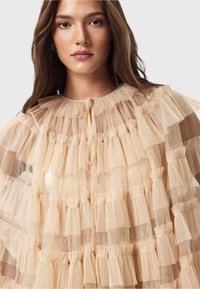 Ljus beige kappa med lager av tylle-volanger, samlade detaljer och rund halsringning. Knytning framtill, genomskinlig struktur, elegant design.