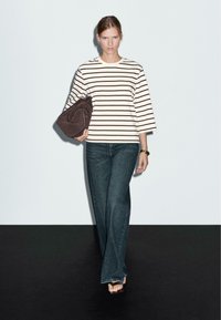 Gestreiftes Hemd mit Dreiviertelärmeln in Creme und Schwarz, kombiniert mit dunkelblauen Schlagjeans. Hält eine braune Wildledertasche in der Hand und steht auf einer dunklen Fläche.
