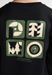 Sweatshirt noir avec quatre blocs graphiques verts présentant des illustrations blanches d'arbres, d'une boussole et de motifs circulaires. Le texte en dessous indique "PETROLIND."