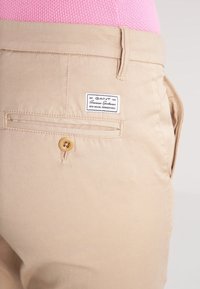 Beige bomullsbukser med en enda bakficka med knappstängning, en märkesskylt och en slät yta.