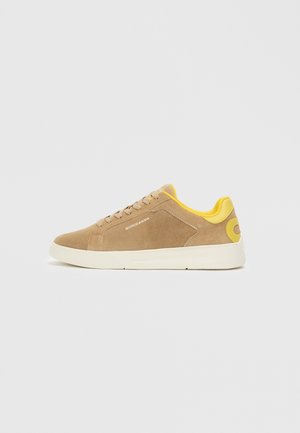 Suede sneakers in light brown with a yellow heel detail, white rubber sole, and beige laces. Logo on the side, textured surface.