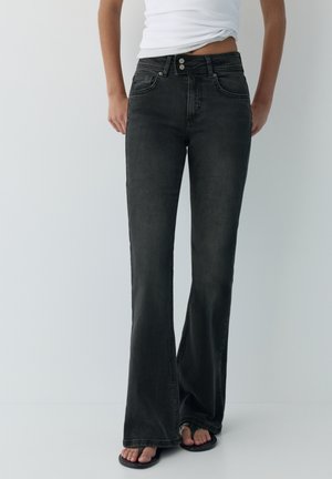 Donna che indossa jeans neri a vita alta a zampa con chiusura a doppio bottone e infradito nere, in piedi davanti a uno sfondo semplice.