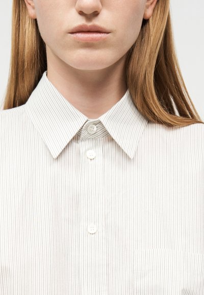 Nahaufnahme einer Person, die ein weißes Hemd mit dünnen vertikalen Streifen und einem geknöpften Kragen trägt, wobei das untere Gesicht und der Hals zu sehen sind.