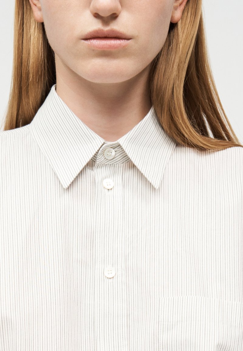 Gros plan d'une personne portant une chemise blanche à fines rayures verticales et un col boutonné, montrant le bas du visage et le cou.