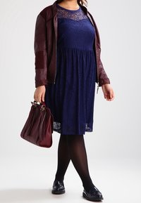 Donna in abito in pizzo blu navy, giacca di pelle bordeaux, collant neri, mocassini neri, che porta una borsa bordeaux, in piedi su uno sfondo bianco.