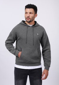 Gray waffle-patterned hoodie with a drawstring hood, front pocket, and small emblem on the chest, paired with black jeans.