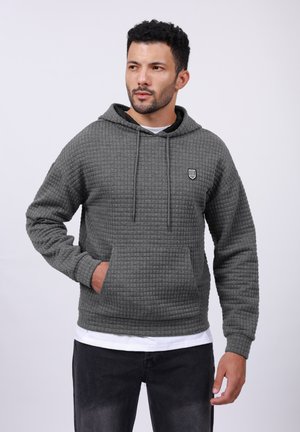 Sudadera gris con patrón de gofre, con capucha ajustable, bolsillo frontal y pequeño emblema en el pecho, combinada con jeans negros.