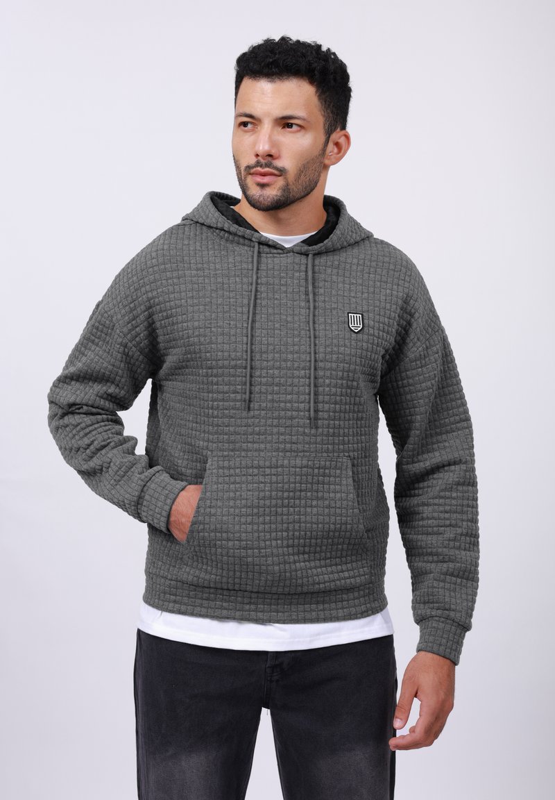 Gray waffle-patterned hoodie with a drawstring hood, front pocket, and small emblem on the chest, paired with black jeans.