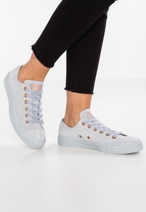 Legs in black cropped pants wearing light gray low-top sneakers with white soles and matching gray laces, standing on white floor.