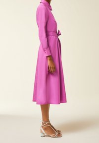 Abito in satin rosa con maniche lunghe, colletto, vita fasciata e gonna a forma A. Abbinato a sandali bianchi con cinturini e tacco a blocco.