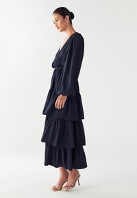 Mornarska plastnata obleka z globokim V-izrezom in dolgimi napihnjenimi rokavi. Ima oprijet pas in tekoče plasti iz lahkotnega blaga, dopolnjena z nude petkami.