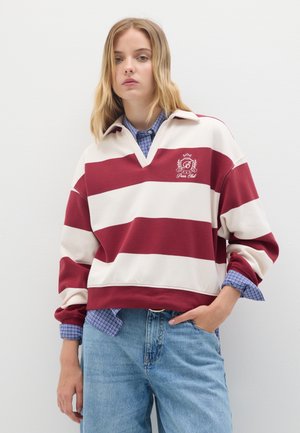 Rot- und cremegestreifter Pullover mit V-Ausschnitt und Kragen. Mit gesticktem Logo auf der Brust. Über einem blauen karierten Hemd getragen.