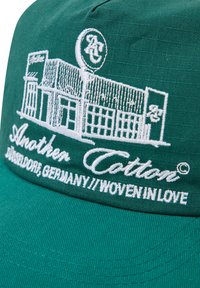 Grüne Baumwollmütze mit weißem besticktem Design eines Gebäudes, Logo und dem Text "Another Cotton, Düsseldorf, Deutschland, Woven in Love."
