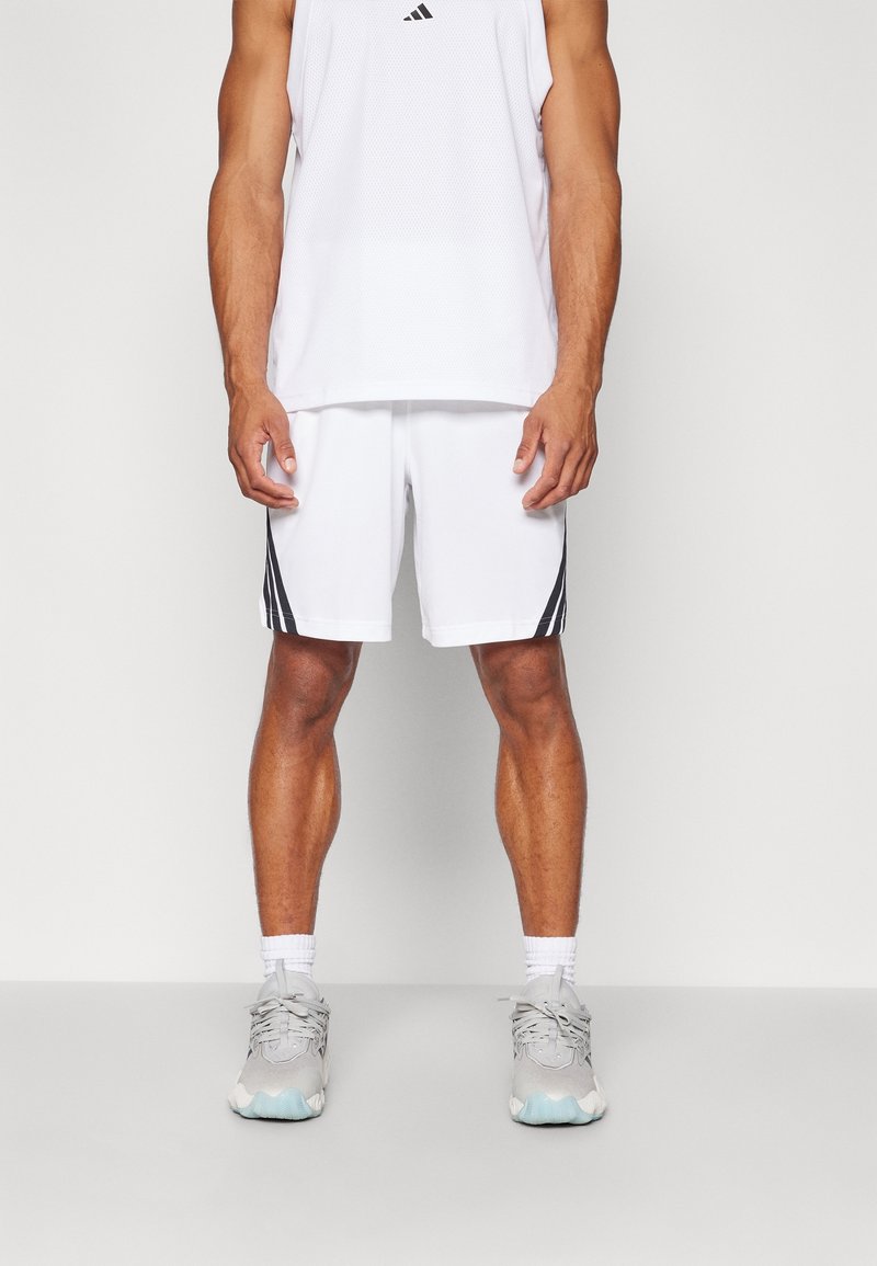 Débardeur de sport blanc Adidas avec logo noir, associé à un short blanc avec des rayures noires sur les côtés, et des baskets grises avec des accents bleu clair.
