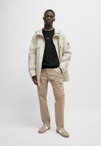 Beige utomhusjacka med två frampocket, över en svart sweatshirt. Okar färgade cargobyxor och beige sneakers med vita detaljer fulländar outfiten.