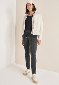 Lichtbeige zip-up jas over een zwart shirt, gecombineerd met donkergrijze joggingbroeken en witte sneakers met lichtblauwe accenten.