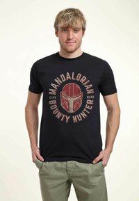 Černé bavlněné tričko s červeným grafickým designem helmy Mandaloriana a textem "Mandalorian Bounty Hunter" v kruhovém uspořádání.