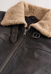 Veste en cuir noir avec col en shearling marron clair, fermeture éclair argentée et détail de sangle à boucle sur le col.