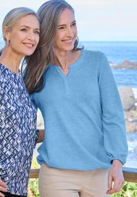 Zwei lächelnde Frauen mittleren Alters stehen nah beieinander, die eine in einem blauen Pullover und beigen Hosen, die andere in einem gemusterten Oberteil, an einer felsigen Küste.