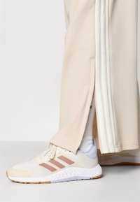 A lábbeli világos krémszínű hálós felsőrésszel, puhán rózsaszín díszítésekkel, fehér, texturált talppal és párnázott nyelvrésszel rendelkezik; beige melegítőnadrággal párosítva.